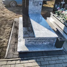 Granitowy nagrobek z inskrypcją i krzyżem, otoczony kostką brukową. Na czarnym tle nagrobka widoczny srebrny krzyż. W tle zaparkowany niebieski samochód.