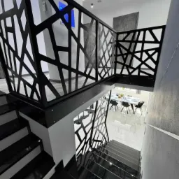 Schody z czarnego kamienia z nowoczesną, ażurową balustradą. Widok z góry na dół, z widocznym fragmentem jadalni w tle. Polerowana powierzchnia stopni odbija światło.