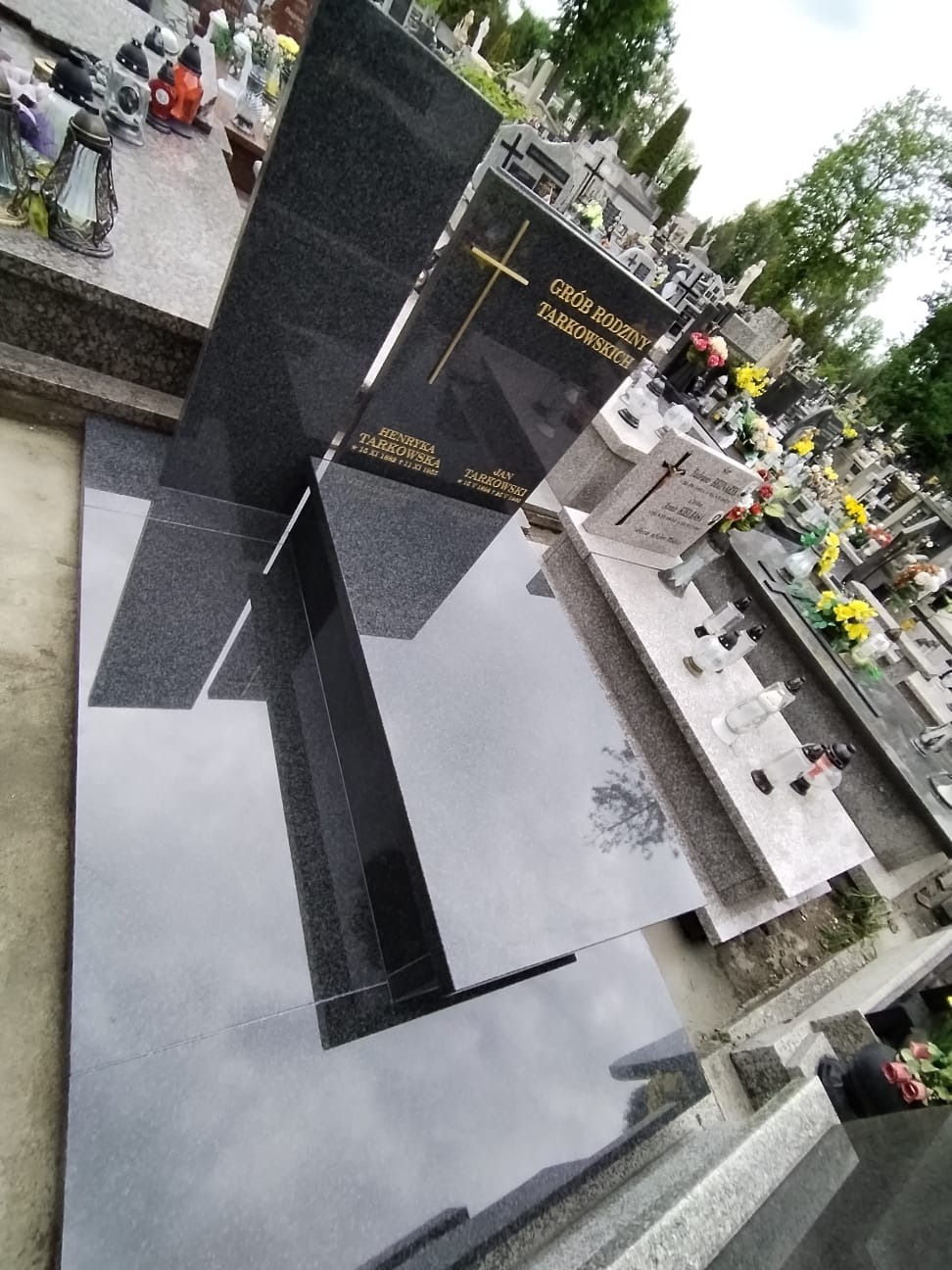 Czarny, nowoczesny nagrobek rodzinny z krzyżem na cmentarzu, z widocznymi refleksami na polerowanej płycie, otoczony innymi nagrobkami i zielenią.