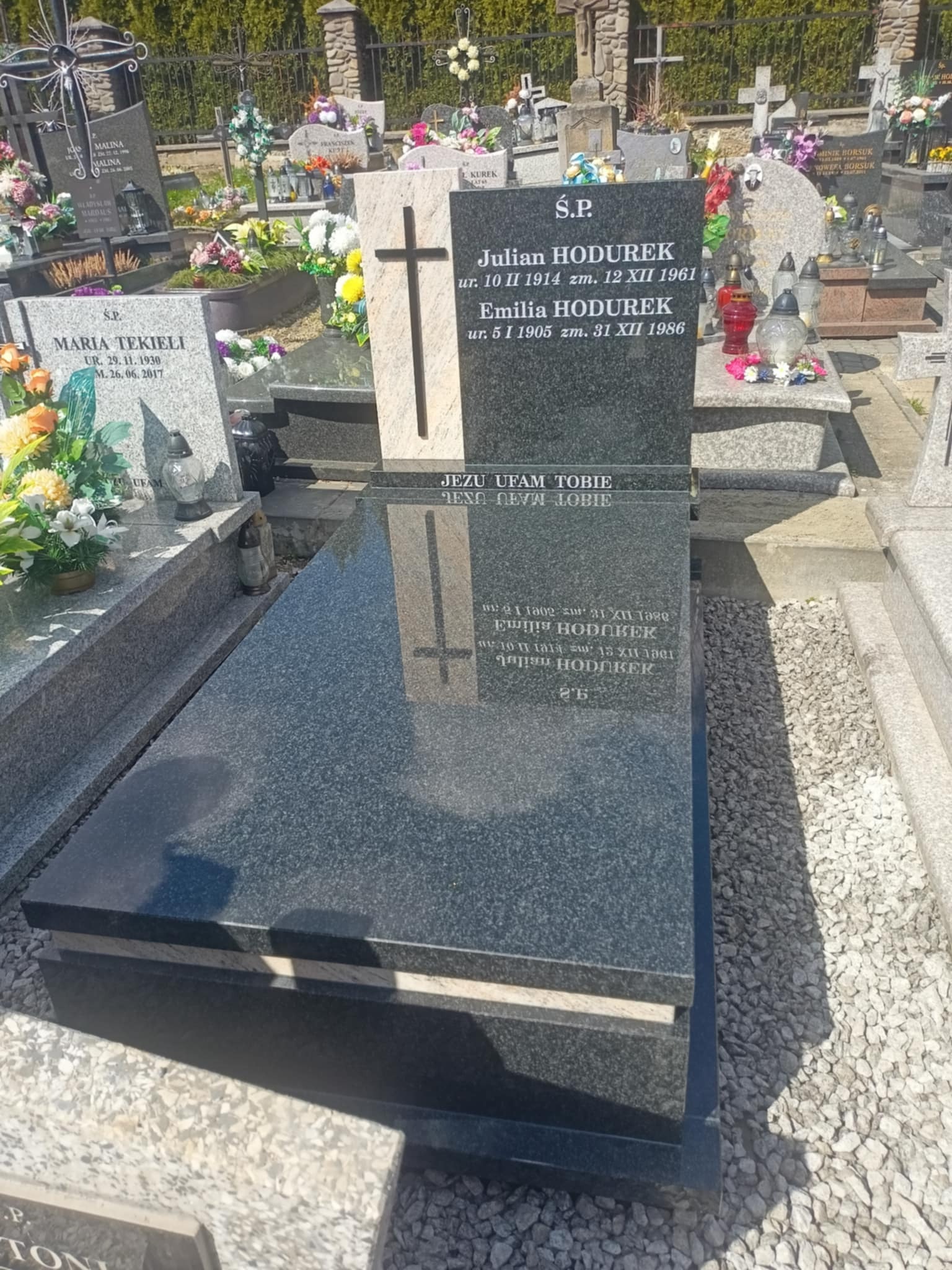 Czarny, polerowany nagrobek z wygrawerowanym krzyżem i inskrypcjami na cmentarzu, otoczony jasnym żwirem i innymi nagrobkami w tle.