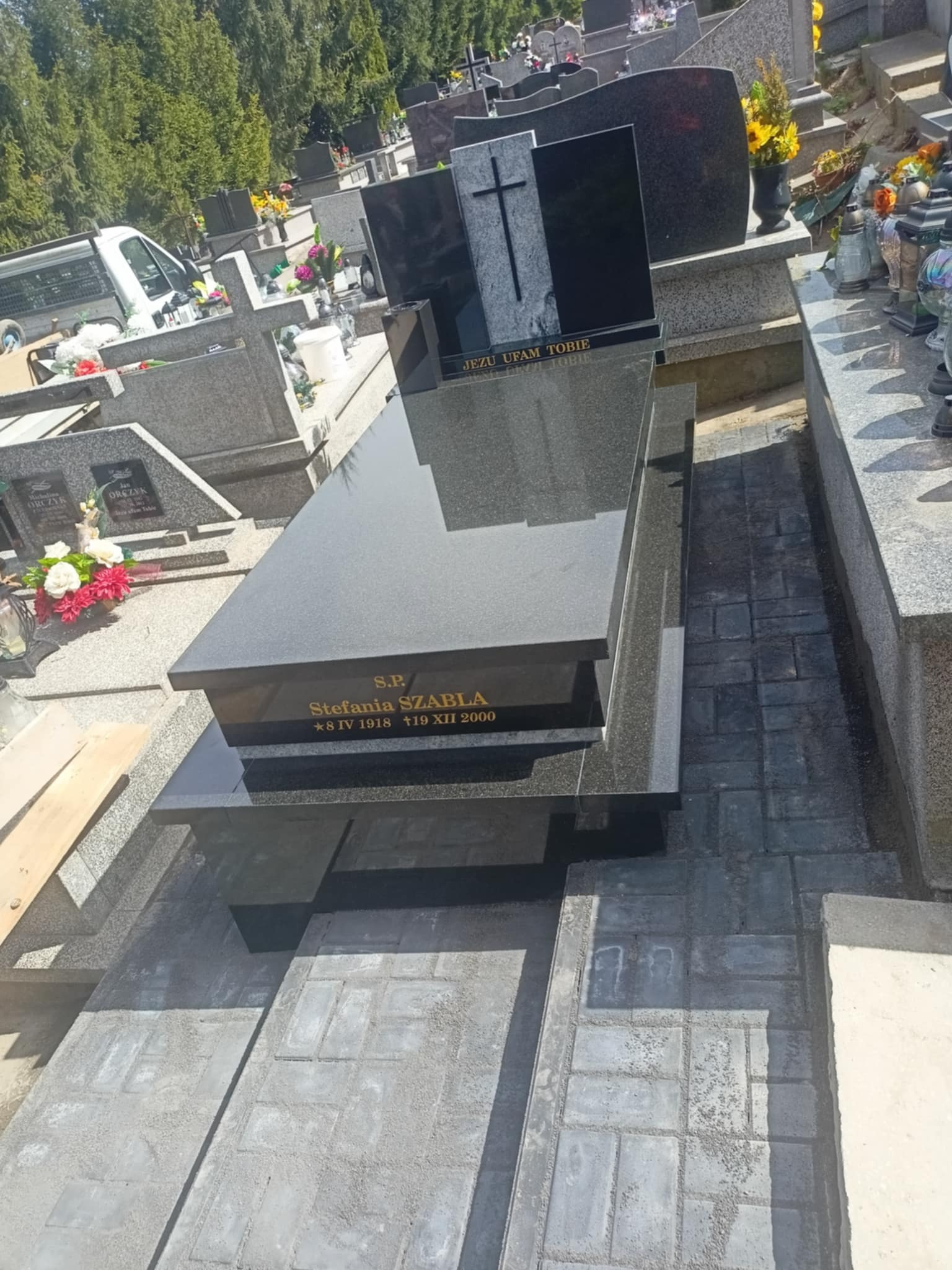 Czarny, nowoczesny nagrobek z wygrawerowanym napisem 'Stefania Szabla' na cmentarzu, otoczony szarą kostką brukową i innymi nagrobkami. Słoneczny dzień.