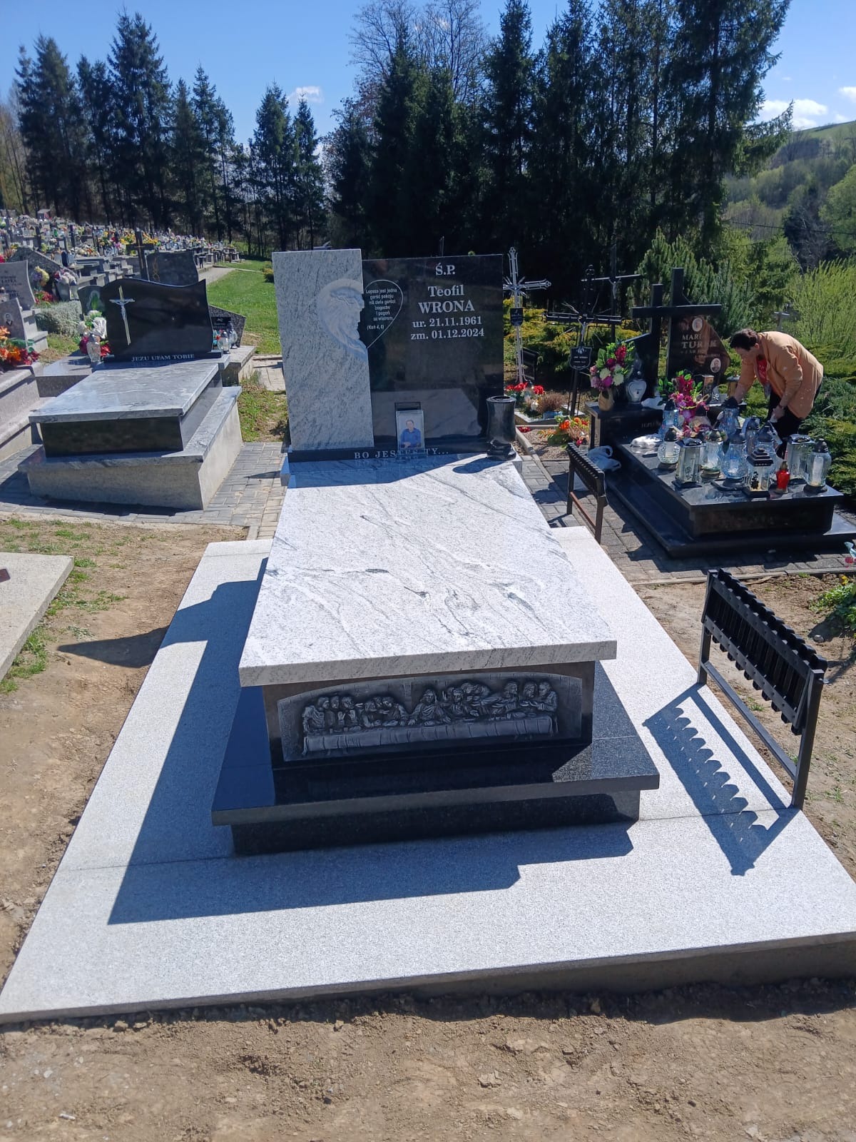 Granitowy nagrobek z płaskorzeźbą Ostatniej Wieczerzy na cmentarzu, w tle drzewa i inne groby. Osoba w oddali. Jasny dzień, błękitne niebo.