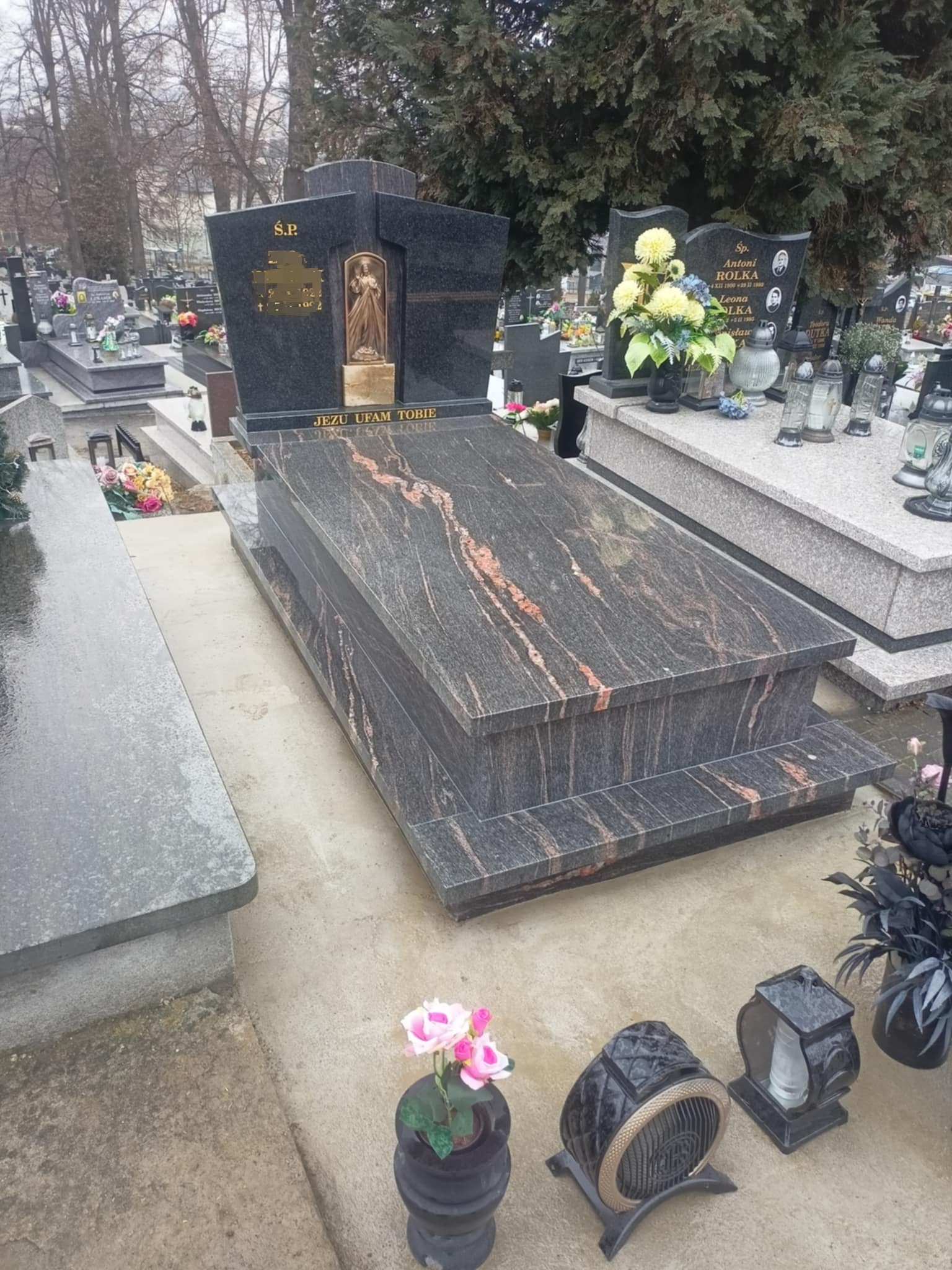 Czarny, granitowy nagrobek z rzeźbą anioła i napisem 'Jezu ufam Tobie' na cmentarzu. Widoczne kwiaty i znicze, pochmurne niebo w tle.