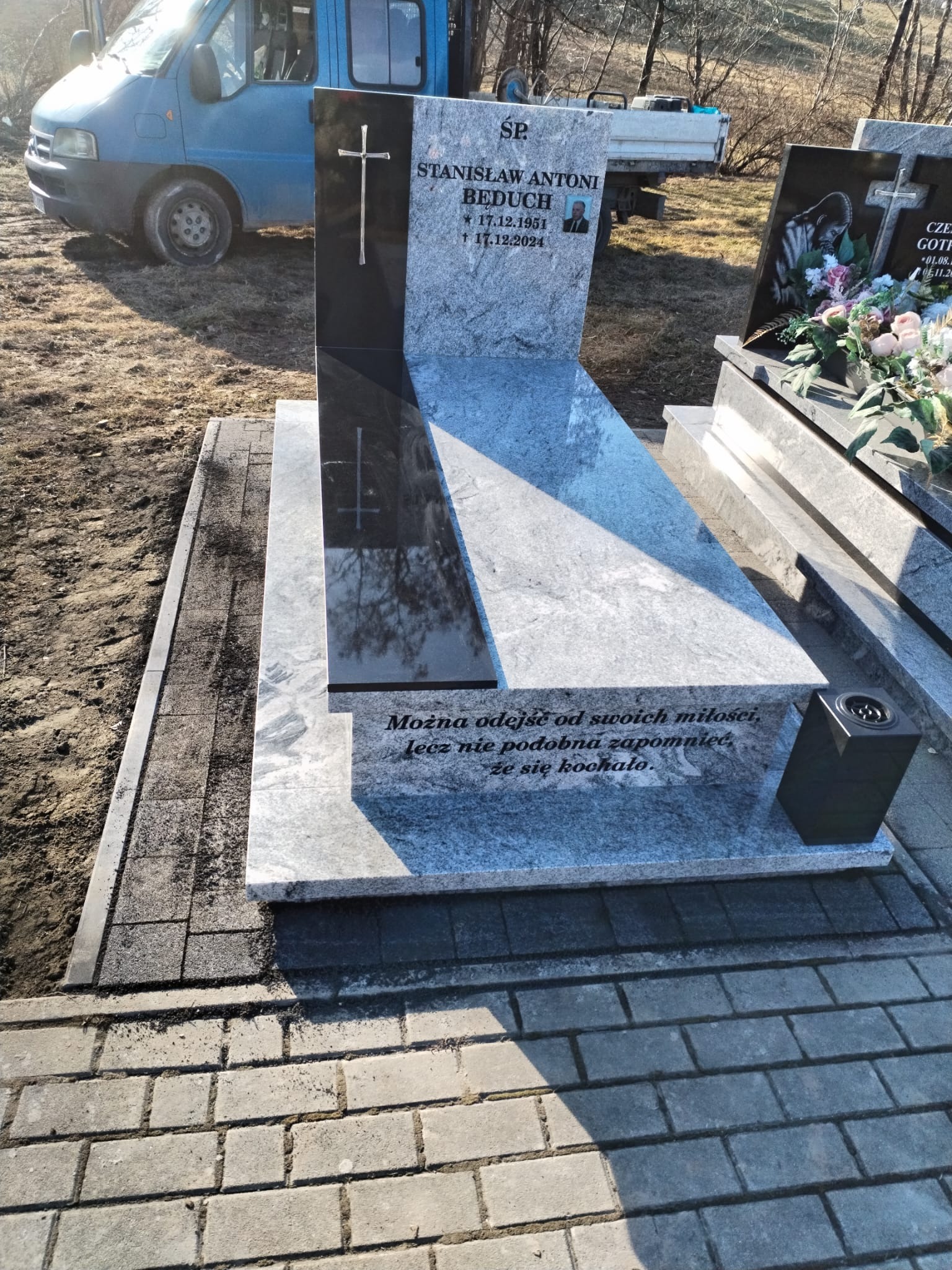 Granitowy nagrobek z inskrypcją i krzyżem, otoczony kostką brukową. Na czarnym tle nagrobka widoczny srebrny krzyż. W tle zaparkowany niebieski samochód.