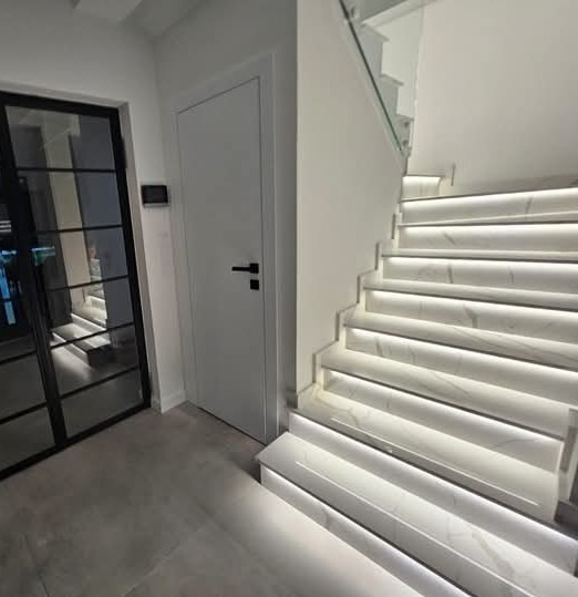 Nowoczesne schody z białego marmuru z podświetleniem LED, szklana balustrada, minimalistyczne drzwi i czarna, przeszklona ściana w tle. Eleganckie wykończenie wnętrza.