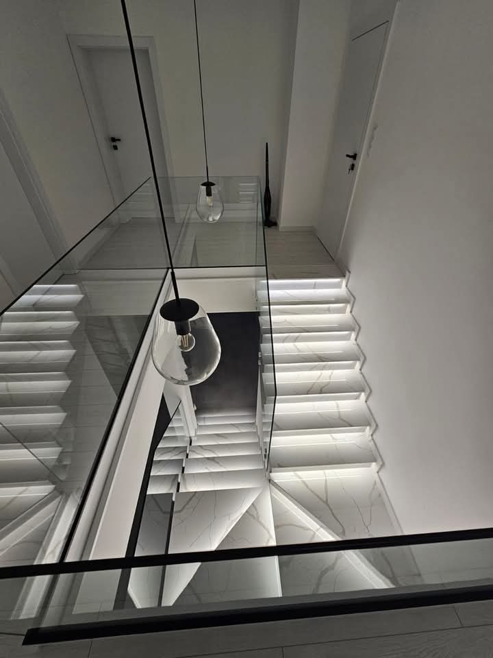 Nowoczesne schody z marmurowymi stopniami, podświetleniem LED i szklaną balustradą, widok z góry z wiszącymi lampami.