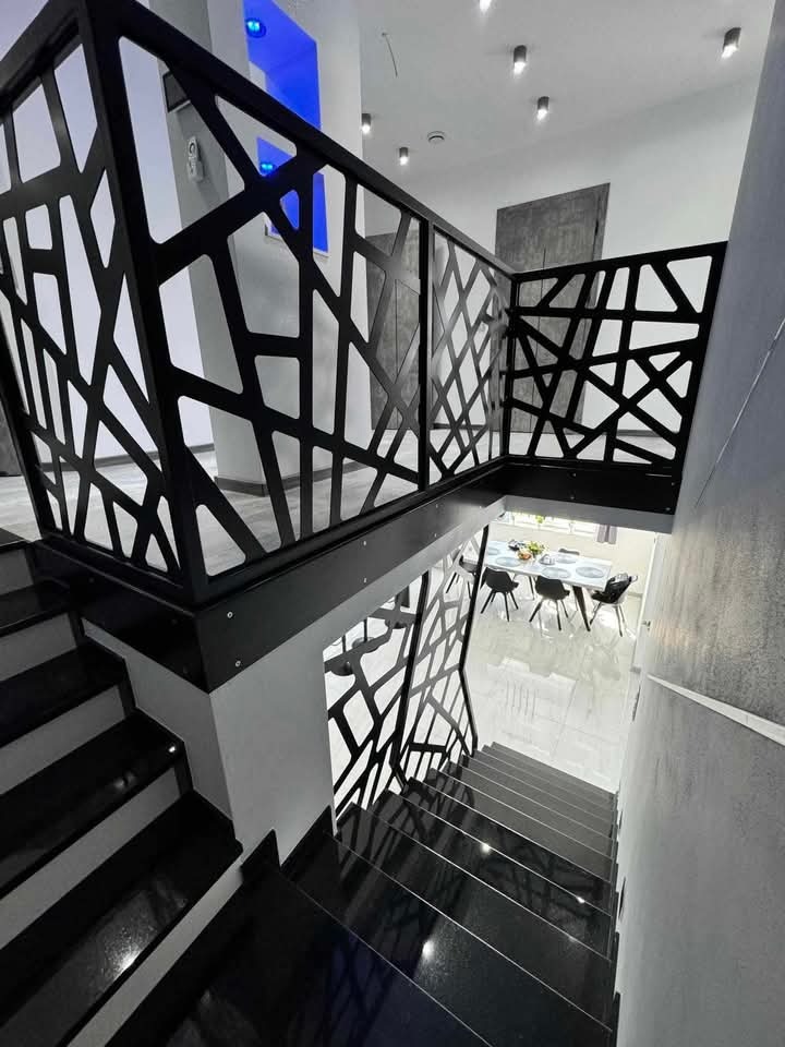 Schody z czarnego kamienia z nowoczesną, ażurową balustradą. Widok z góry na dół, z widocznym fragmentem jadalni w tle. Polerowana powierzchnia stopni odbija światło.