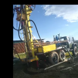 Żółta wiertnica zamontowana na ciężarówce, używana do wiercenia studni w Gorlicach, z widoczną wyżłobioną ziemią wokół miejsca wiercenia i niebieskim niebem w tle.