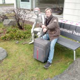 Usługi remontowo-budowlane Krzysztof Popławski - Mężczyzna w kurtce i jeansach, stojący obok srebrnego manekina i walizki na kółkach, na tle budynku z oknami i ławką z napisem.