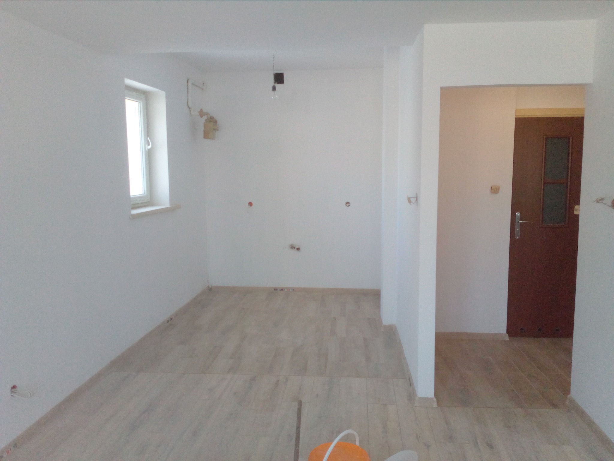 Wnętrze pokoju po remoncie z położoną podłogą imitującą drewno, widoczne ślady po instalacji elektrycznej na ścianach, surowa żarówka zwisająca z sufitu i pomarańczowe wiadro w rogu.