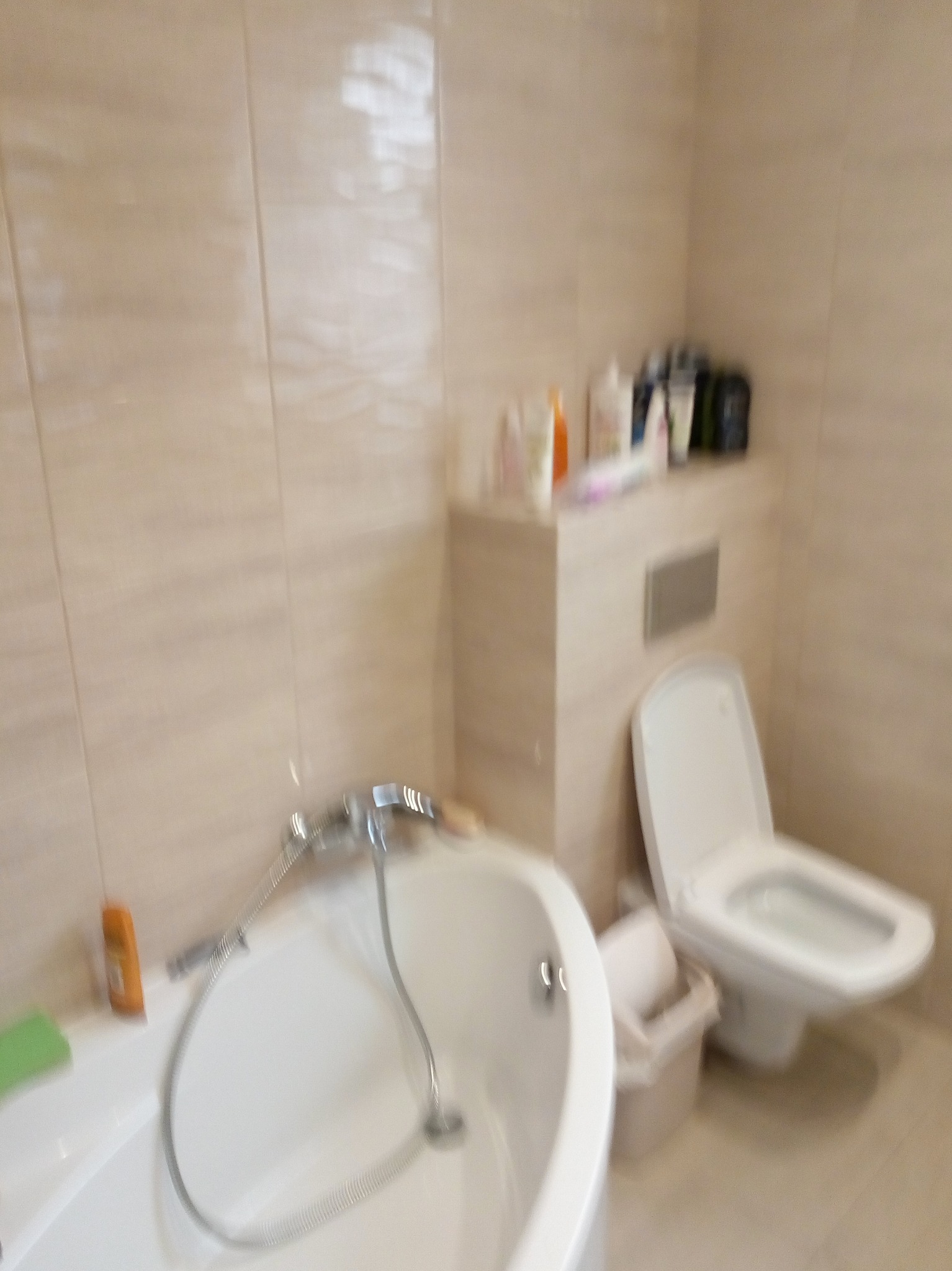 Wykończona łazienka z wanną, toaletą i beżowymi płytkami na ścianach, z widocznymi kosmetykami na półce.