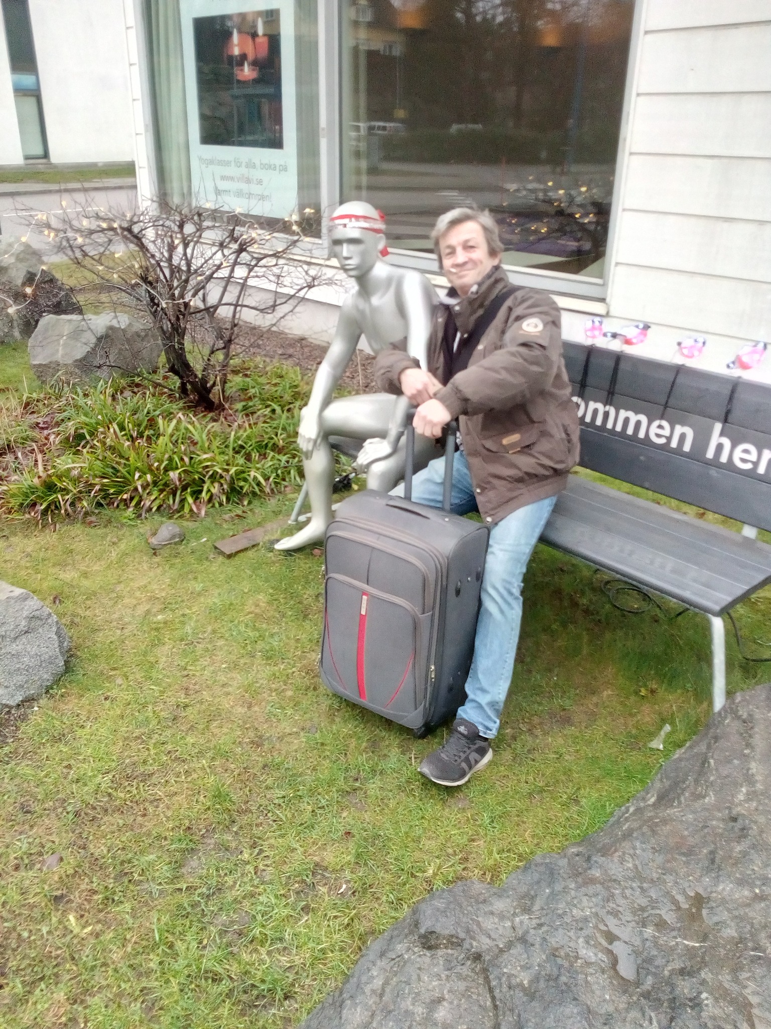 Mężczyzna w kurtce i jeansach, stojący obok srebrnego manekina i walizki na kółkach, na tle budynku z oknami i ławką z napisem.