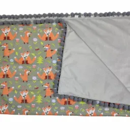 Dziecięcy kocyk z motywem lisów na szarym tle, wykończony szarymi pomponami, ukazujący gładką, szarą podszewkę.