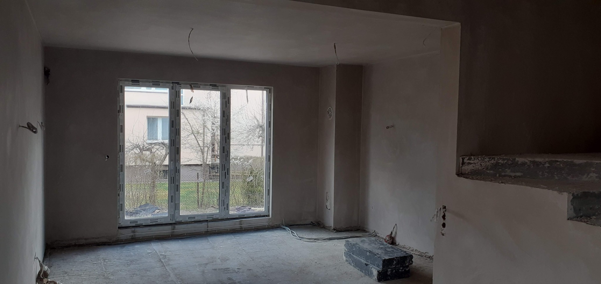 Wnętrze po tynkowaniu maszynowym: gładkie ściany w surowym stanie, duże okno z widokiem na zewnątrz, przewody elektryczne i skrzynka na podłodze.