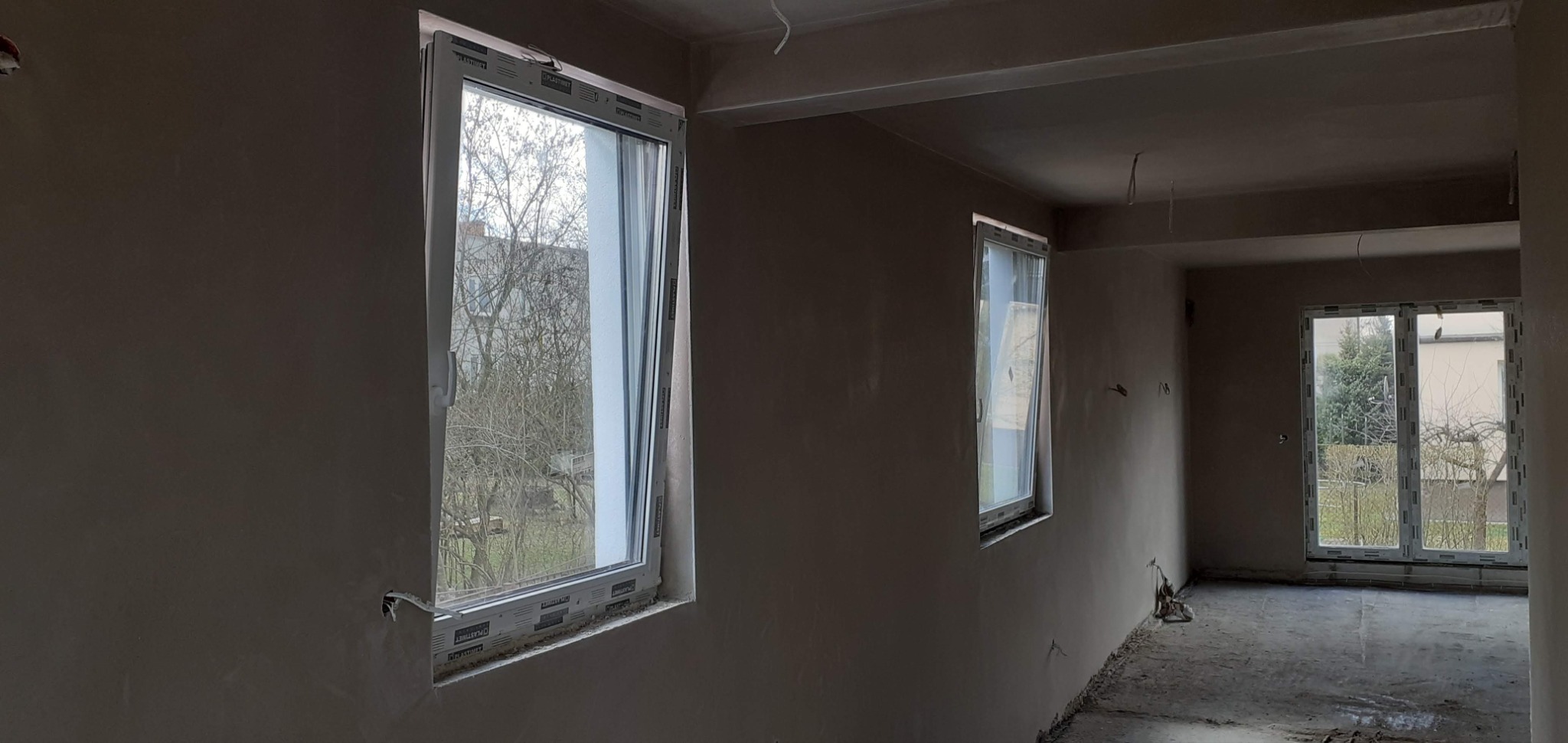 Wnętrze po nałożeniu gładzi: ściany i sufit pokryte świeżym tynkiem, widoczne okna z widokiem na zewnątrz. Detal: wystające przewody elektryczne.