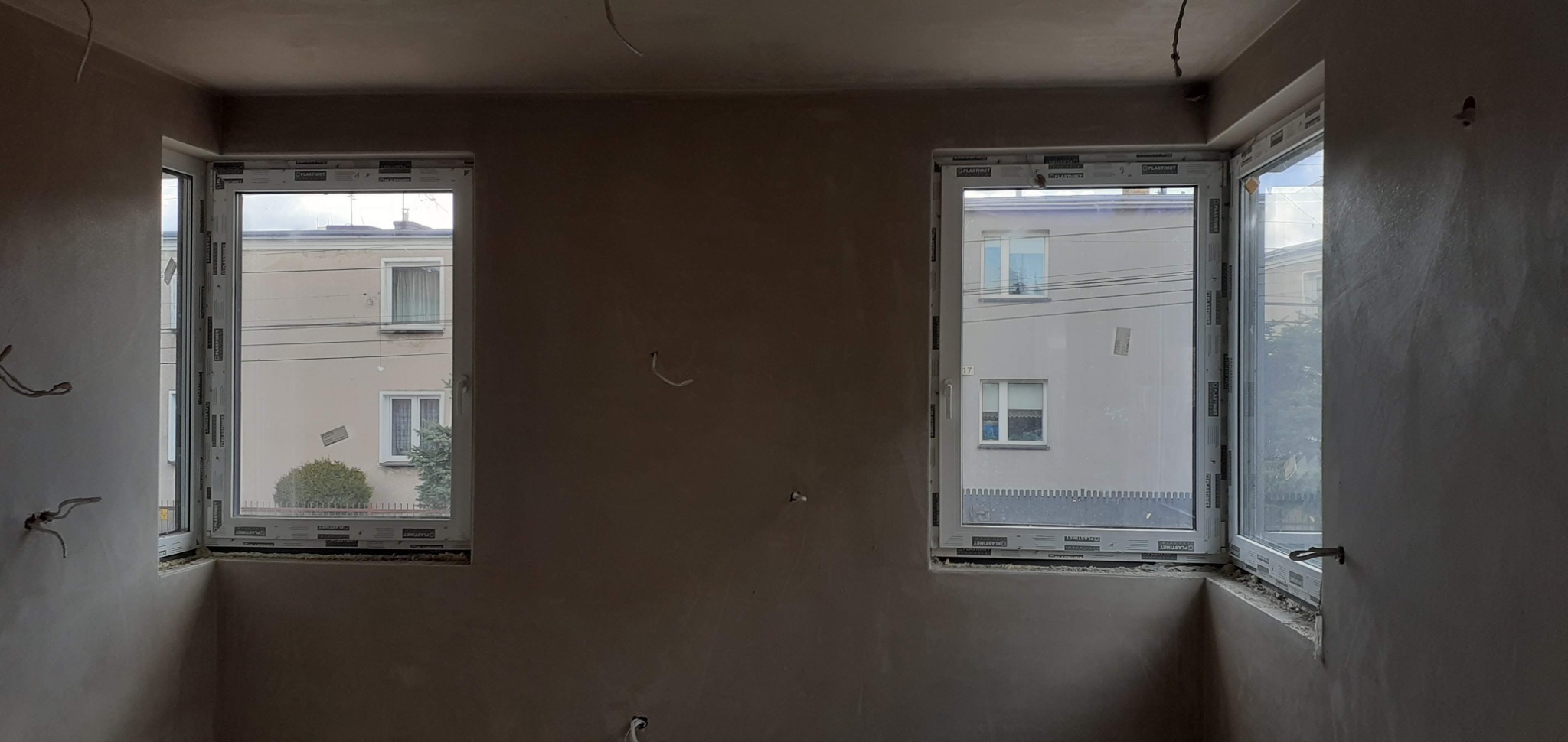 Pokój po tynkowaniu maszynowym, widoczne dwa okna z widokiem na budynek. Na ścianach szare, świeże tynki. Widoczne wystające kable elektryczne.
