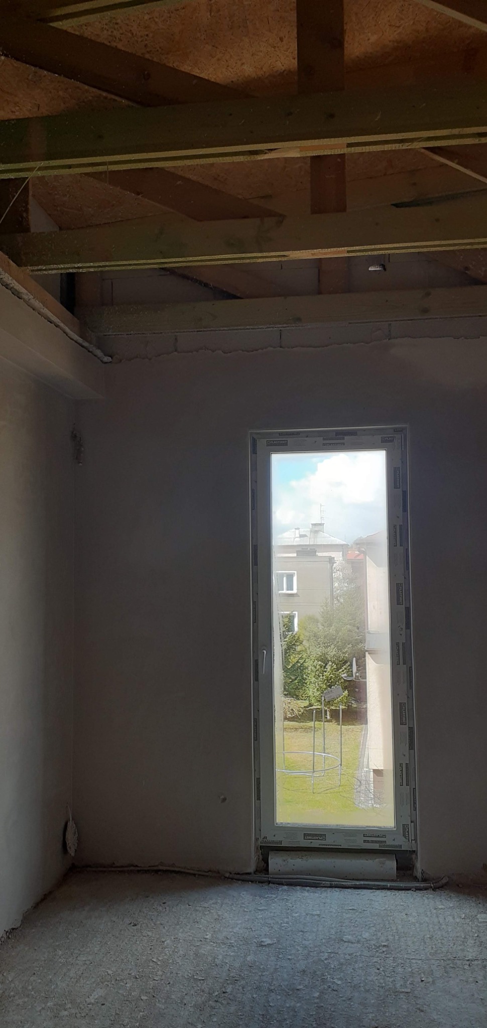 Wnętrze pokoju w stanie surowym, ściany po nałożeniu tynku, widoczne drewniane elementy konstrukcji sufitu i okno z widokiem na zewnątrz.