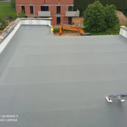 Nowy, płaski dach pokryty szarą membraną, z widoczną izolacją na krawędziach i koparką JCB w tle, na tle niedokończonego budynku z czerwonej cegły.