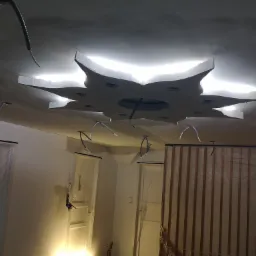 Niedokończony sufit podwieszany w kształcie gwiazdy z podświetleniem, widoczne kable i instalacje elektryczne, zabezpieczone drzwi folią malarską.