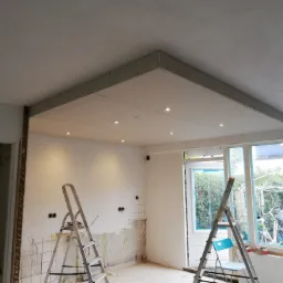 Sufit podwieszany z płyt kartonowo-gipsowych z oświetleniem punktowym w trakcie montażu, widoczne drabiny i ślady po zdjętych płytkach na ścianie.