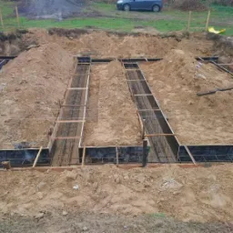 Przygotowanie szalunku i zbrojenia pod fundamenty domu jednorodzinnego, widoczne deski szalunkowe, stalowe pręty zbrojeniowe i wykop z ziemią, w tle trawa i samochód.