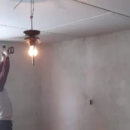 Pracownik w trakcie montażu oświetlenia w pomieszczeniu z surowymi ścianami i sufitem z płyt kartonowo-gipsowych; widoczne narzędzia i przewody.