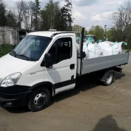 Biały samoch&oacute;d dostawczy Iveco z otwartą skrzynią ładunkową, wypełnioną dużymi workami z jasnym materiałem, prawdopodobnie ziemią lub żwirem, zaparkowany na uboczu drogi gruntowej w słoneczny dzień.