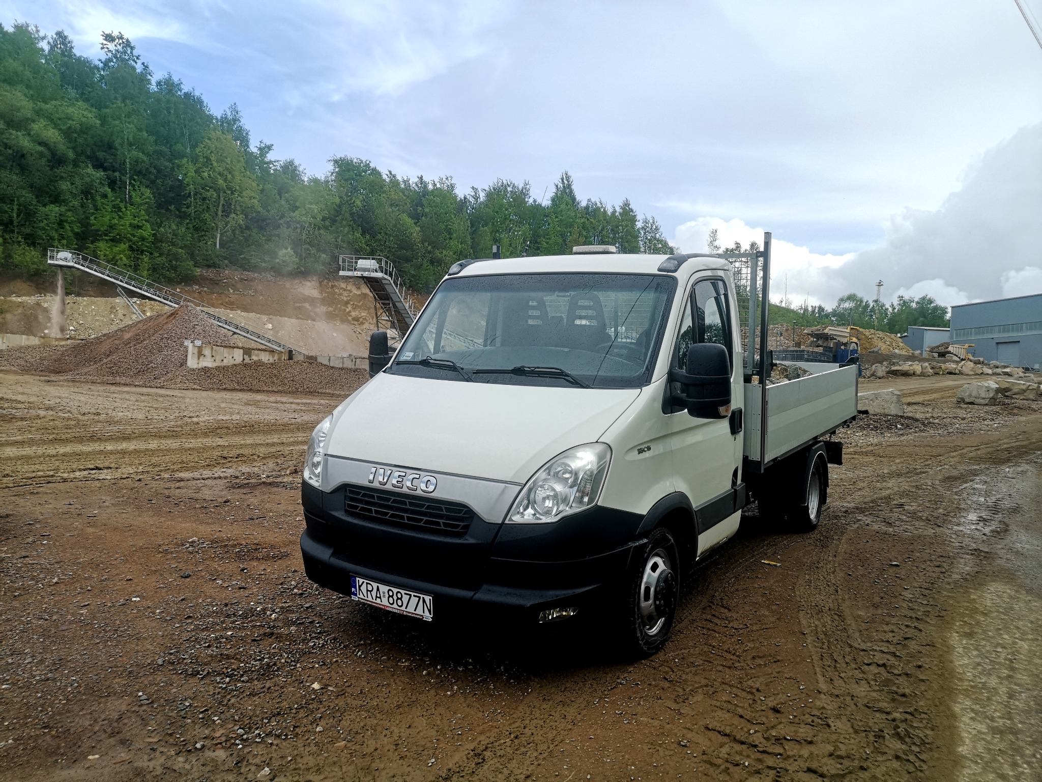 Biała ciężarówka Iveco z otwartą skrzynią ładunkową na placu budowy, w tle hałda materiału skalnego i las.
