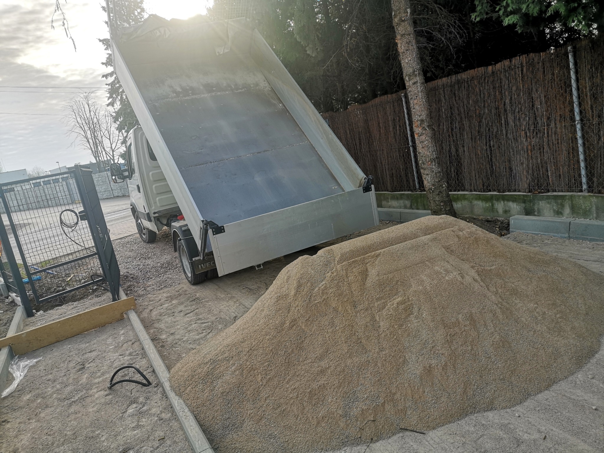 Wyładowany z wywrotki kopiec drobnego kruszywa na placu budowy, widoczne krawężniki i ogrodzenie z siatki.