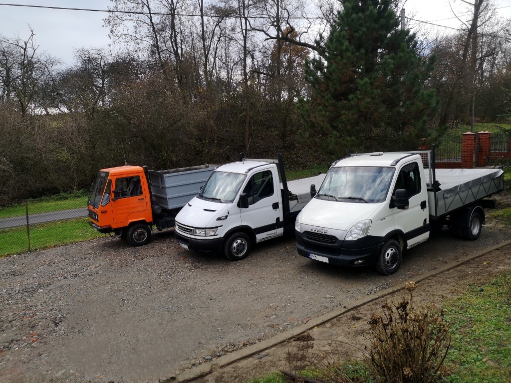 Trzy samochody dostawcze: pomarańczowy, biały i biały, zaparkowane na żwirowym podłożu, z drzewami w tle.