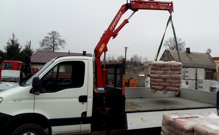 Ciężarówka z dźwigiem Ferrari 530 podnosi paletę z workami, prawdopodobnie cementu lub zaprawy, na tle budynków i pochmurnego nieba.