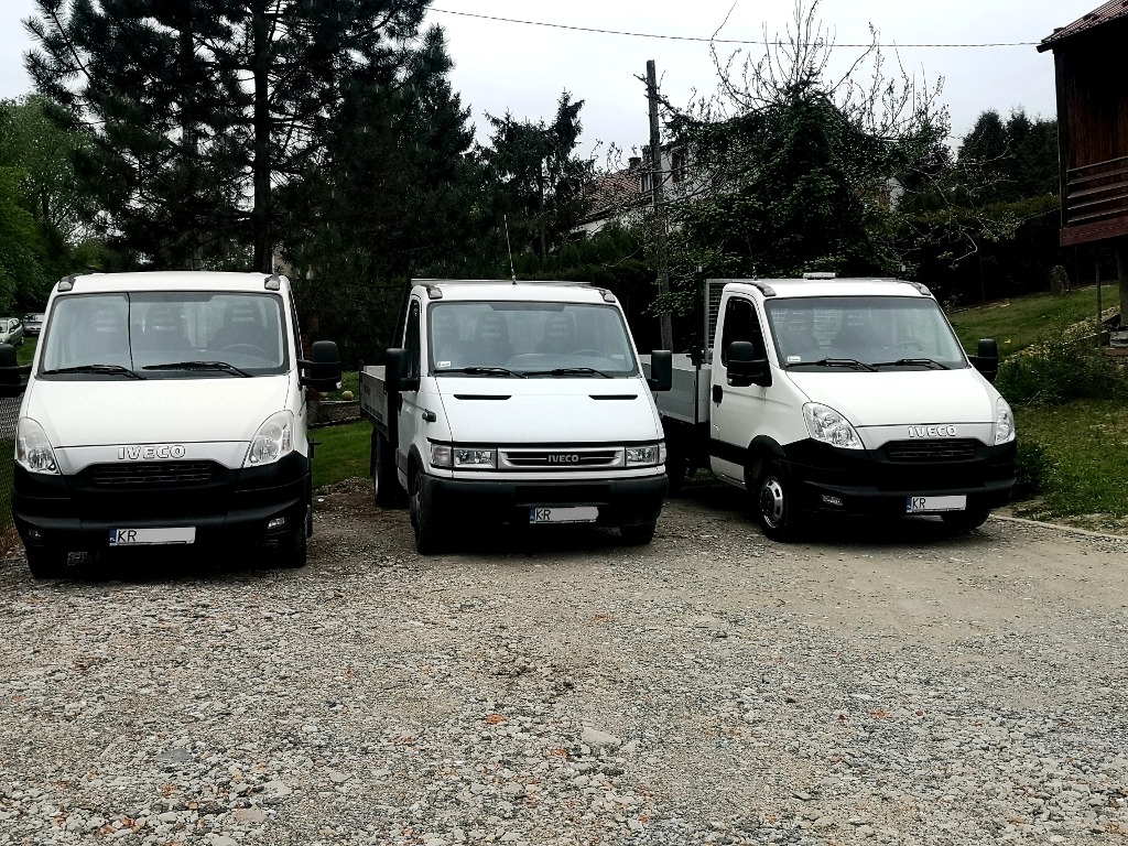 Trzy białe samochody dostawcze Iveco zaparkowane na żwirowym placu, widok z przodu, w tle zieleń drzew i budynki mieszkalne.