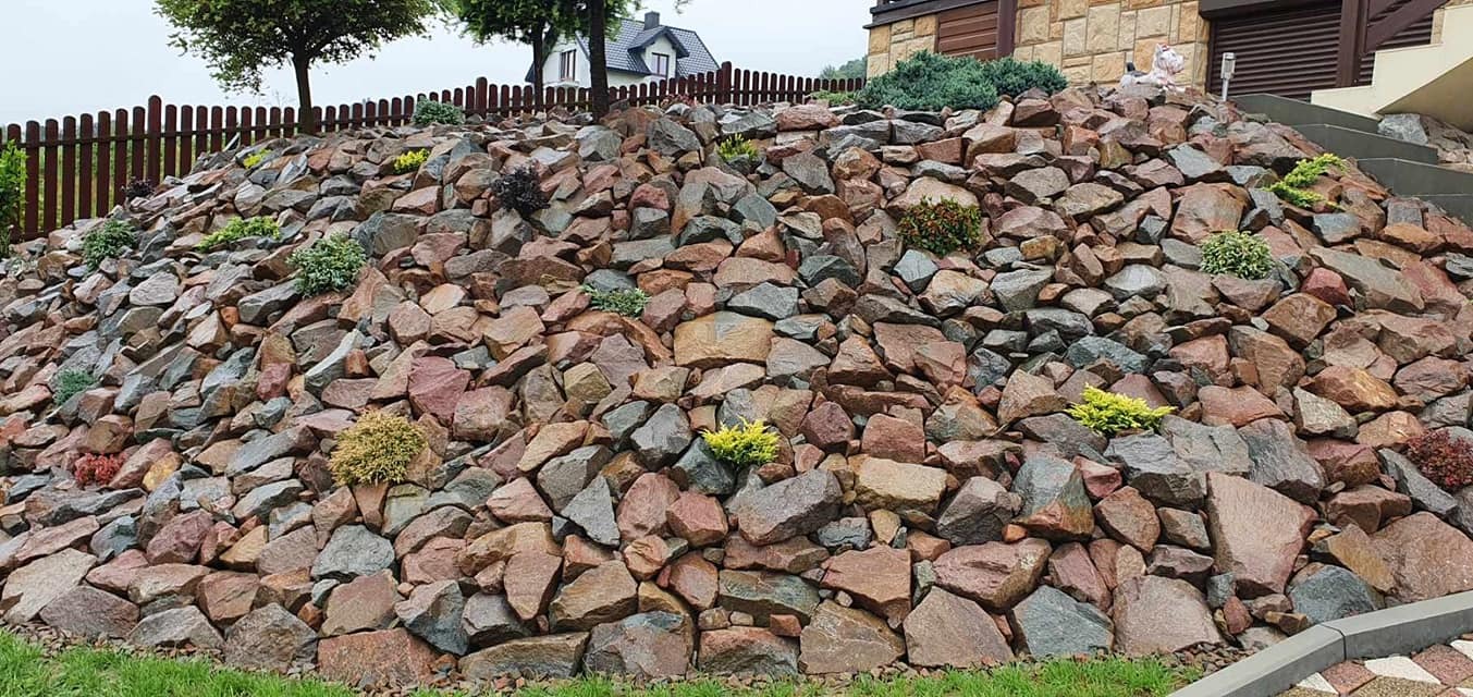 Skarpa wzmocniona ułożonymi kamieniami różnej wielkości i koloru, z nasadzonymi niskimi krzewami iglastymi i liściastymi, w tle drewniany płot i dom.