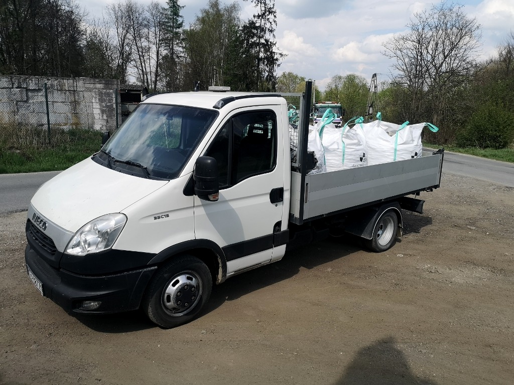 Biały samochód dostawczy Iveco z otwartą skrzynią ładunkową, wypełnioną dużymi workami z jasnym materiałem, prawdopodobnie ziemią lub żwirem, zaparkowany na uboczu drogi gruntowej w słoneczny dzień.