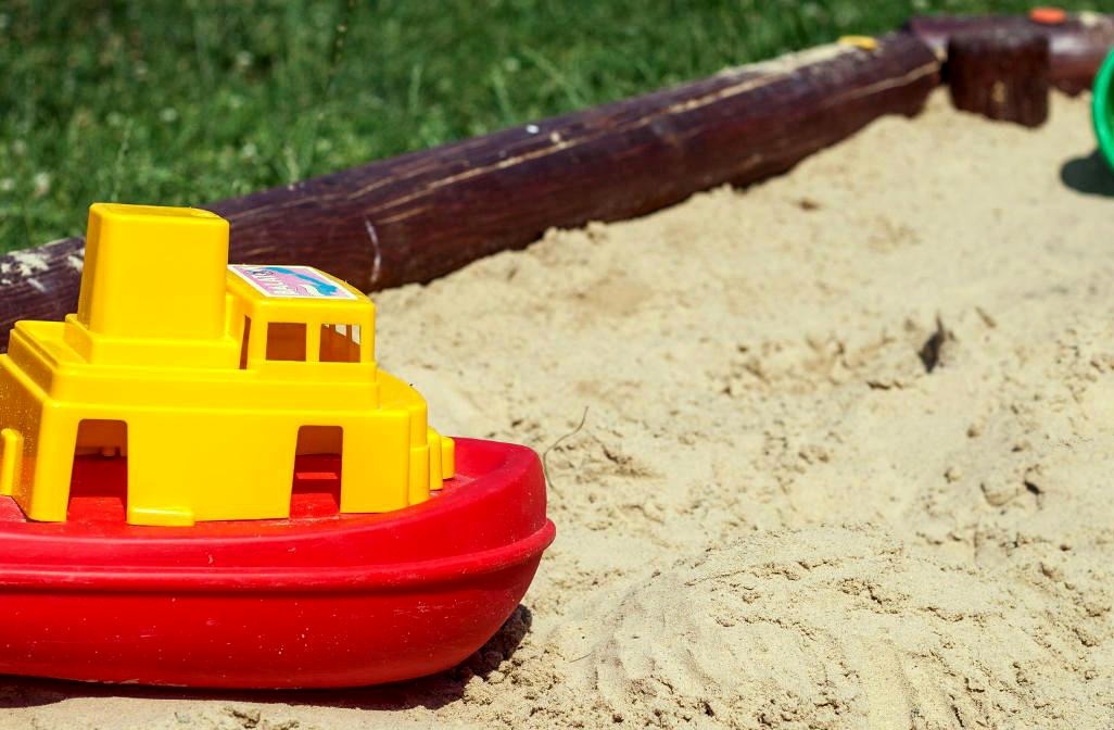 Plastikowa, czerwono-żółta zabawka - statek w piaskownicy z drewnianym obrzeżem, prawdopodobnie w Krakowie.