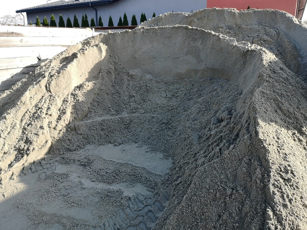 Duża pryzma piasku budowlanego z wyraźnymi śladami opon pojazdu, w tle widoczny fragment budynku mieszkalnego i rząd tuj.