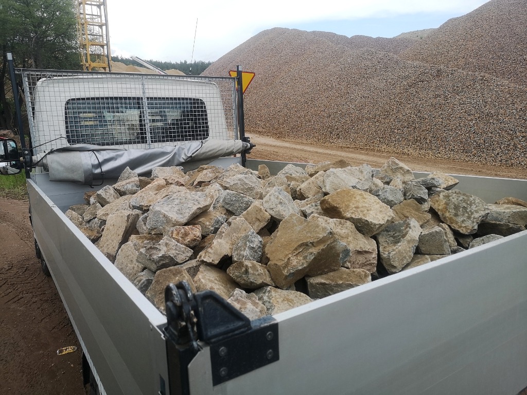 Ładunek kamieni w skrzyni samochodu dostawczego, w tle hałda kruszywa i żółty żuraw budowlany.
