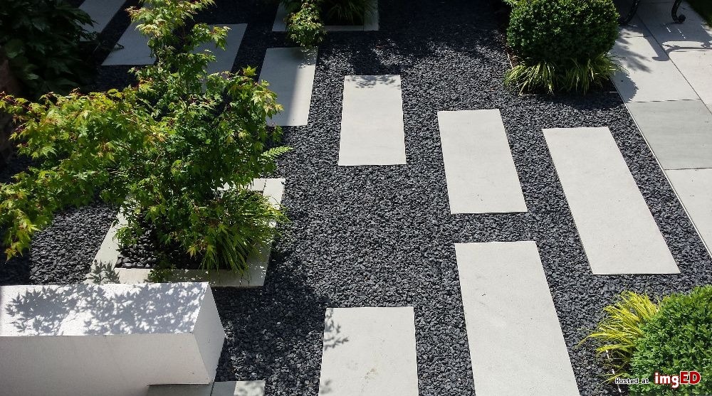 Nowoczesny ogród z ścieżką z betonowych płyt otoczonych ciemnym drobnym kamieniem, z nasadzeniami roślin ozdobnych w geometrycznych donicach.