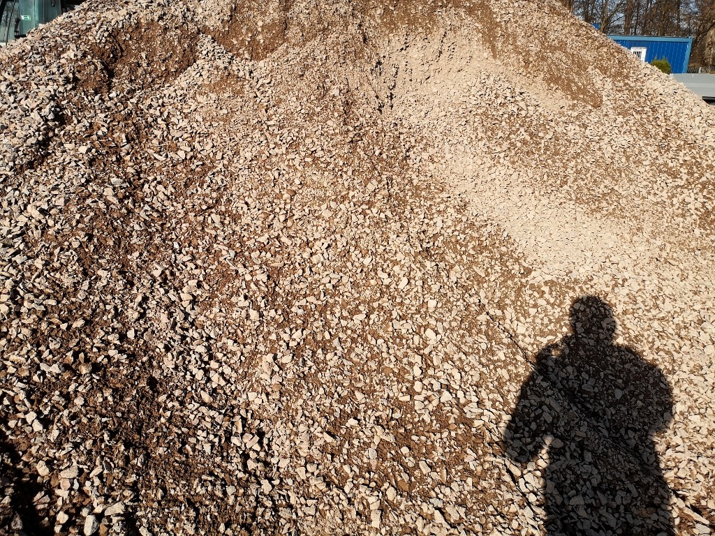 Duża pryzma jasnego żwiru z widocznym cieniem osoby robiącej zdjęcie, na tle częściowo widoczny budynek.