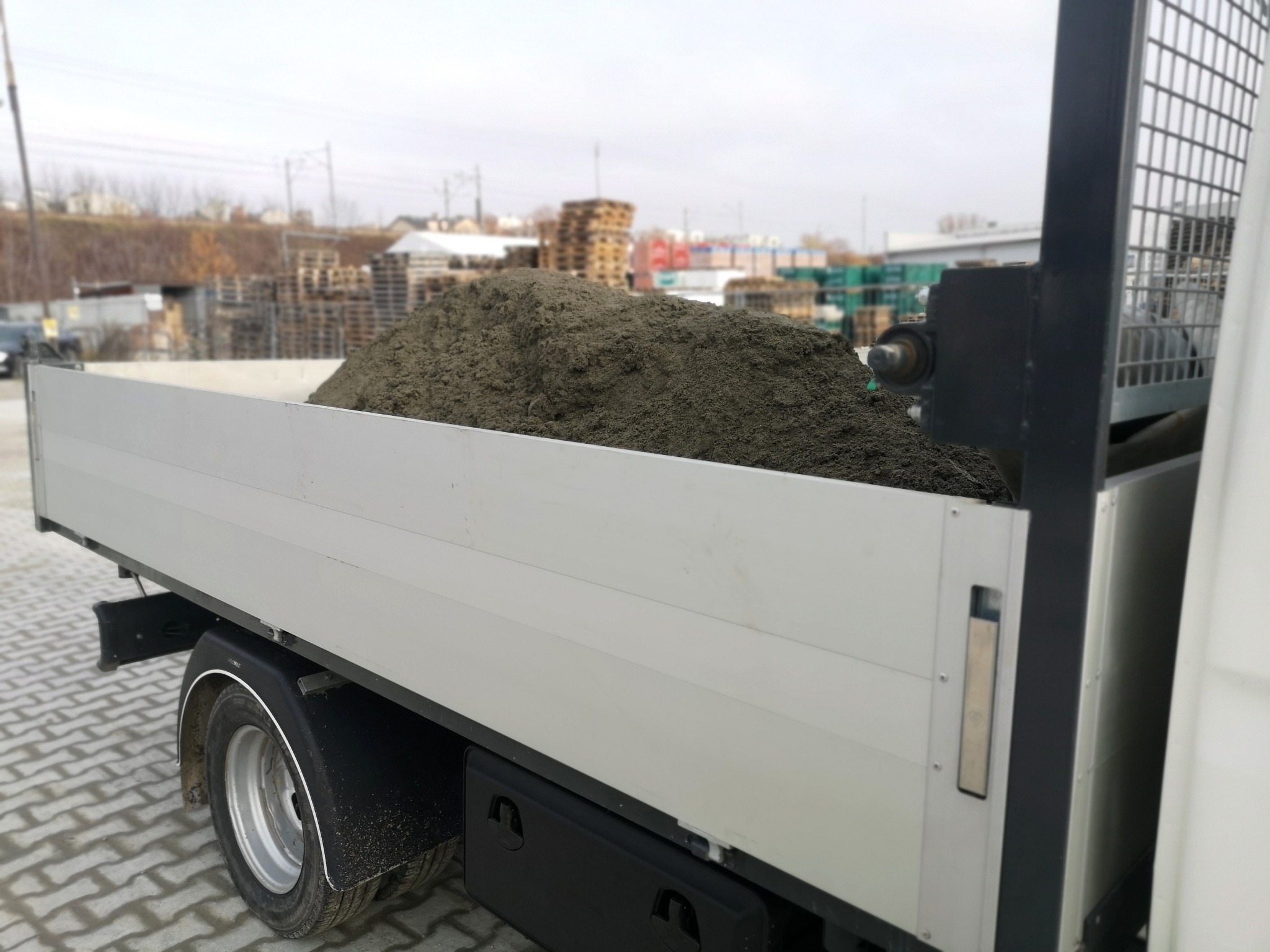 Ciężarówka z platformą załadowana stertą ciemnego kruszywa na tle składu materiałów budowlanych.