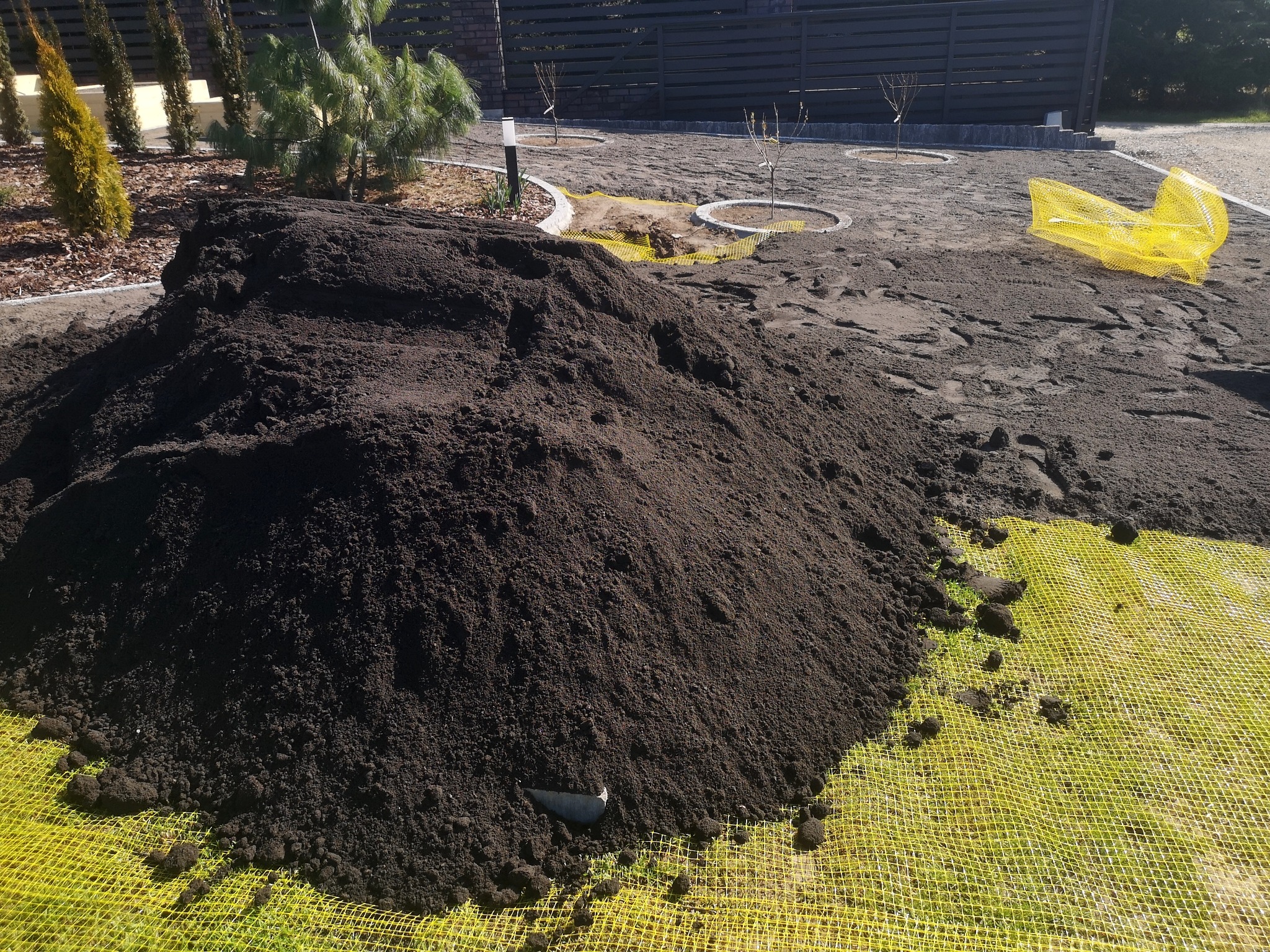 Kupa ciemnej ziemi ogrodowej na żółtej siatce w ogrodzie, widoczne młode drzewka i elementy małej architektury.