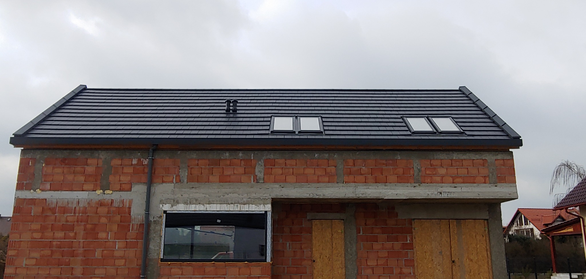 Dom w trakcie budowy z czarnym dachem z dachówki ceramicznej, trzema oknami dachowymi i kominkiem wentylacyjnym na tle pochmurnego nieba.