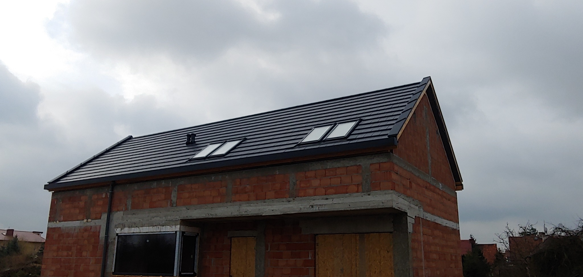 Dom w trakcie budowy z nowym, czarnym dachem z dachówki ceramicznej i dwoma oknami dachowymi, na tle pochmurnego nieba.
