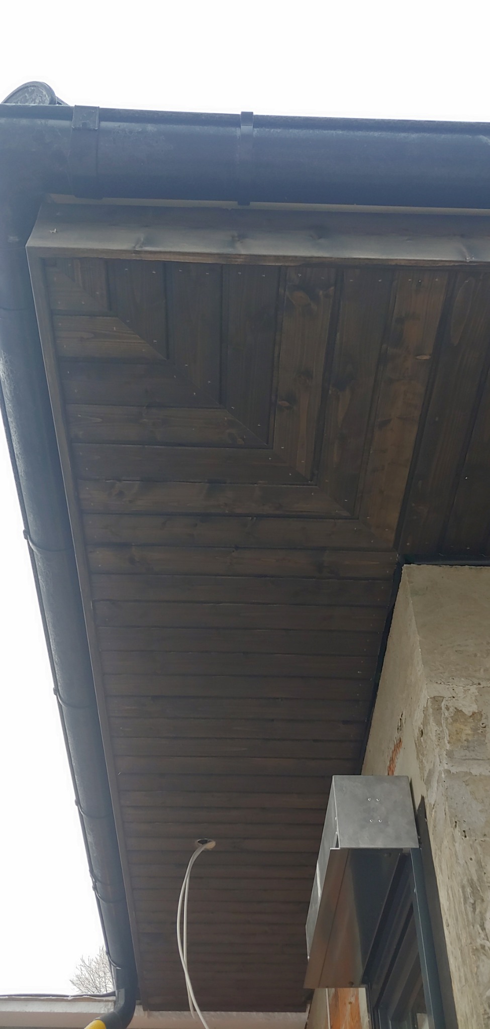 Podbitka dachowa z ciemnego drewna z widocznymi gwoździami, rynna i fragment ściany budynku.