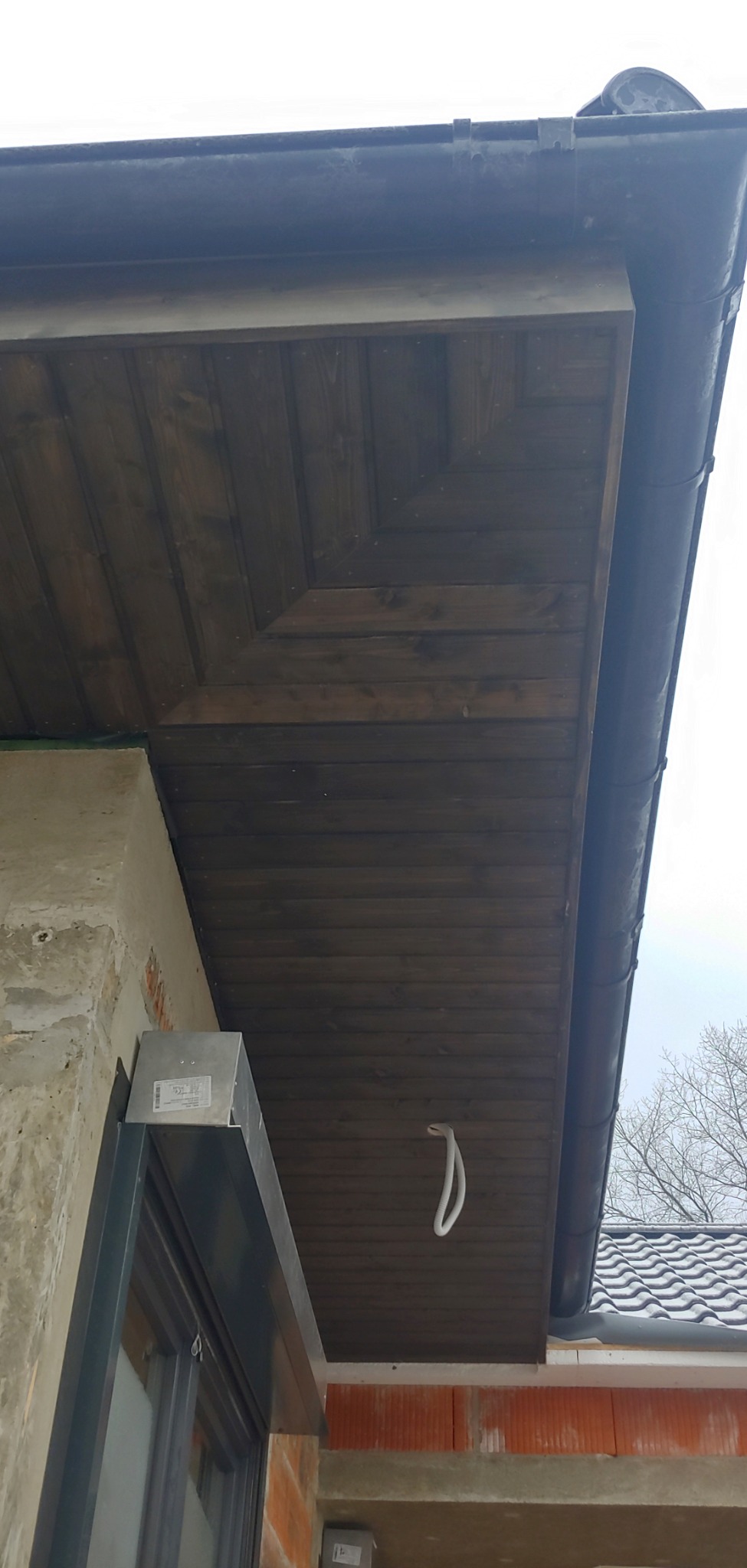 Drewniana podbitka dachowa ułożona w jodełkę, z widoczną rynną i fragmentem elewacji w trakcie budowy.