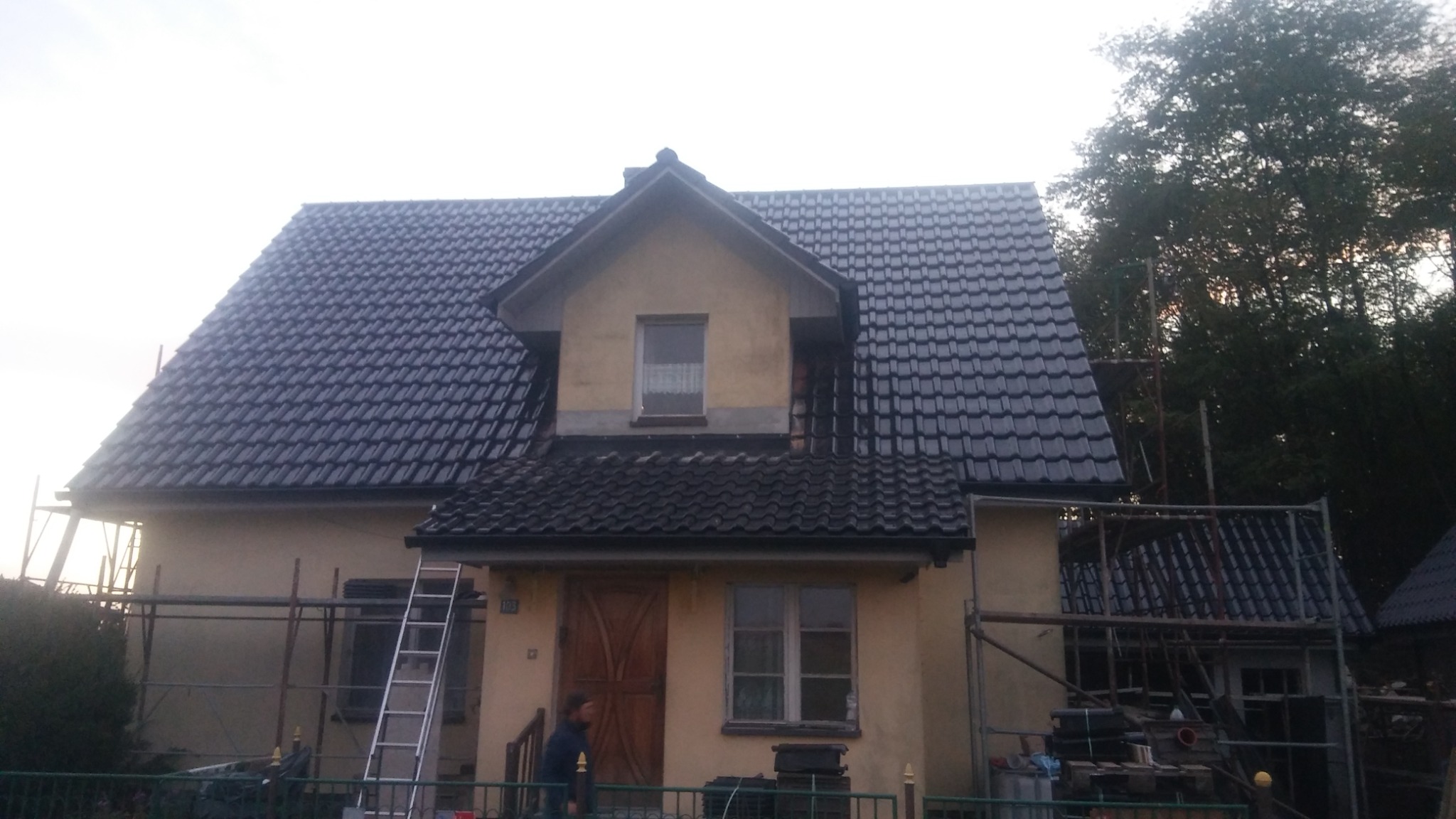 Dom jednorodzinny z nowym, błyszczącym, czarnym dachem z dachówki ceramicznej, widoczne rusztowania i drabina, sugerujące prace budowlane w toku.