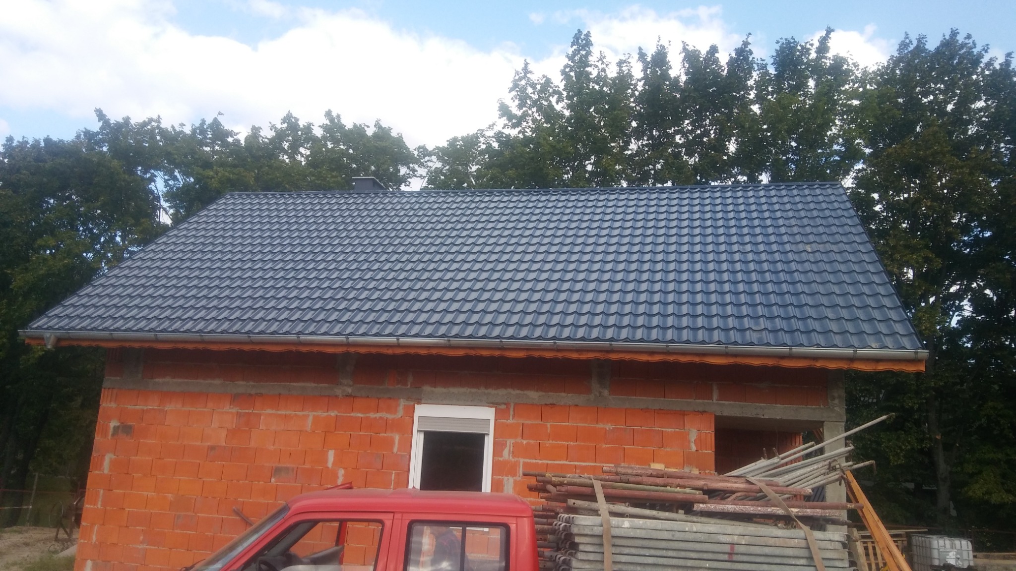 Nowy, ciemnoszary dach z blachodachówki na budynku w trakcie budowy, widoczne rusztowania i czerwony samochód.