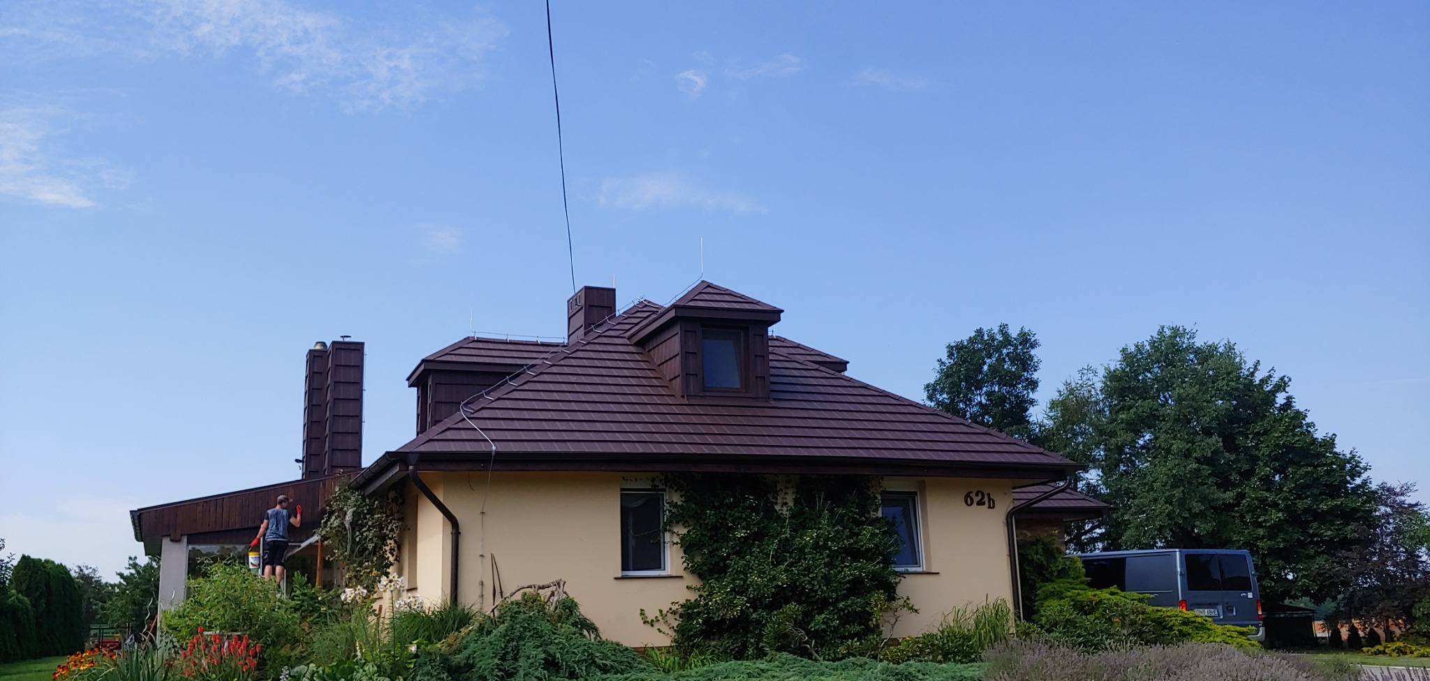 Pracownik na drabinie przy dachu domu w Nysie, widoczne prace malarskie przy kominie, brązowa dachówka i kremowa elewacja.