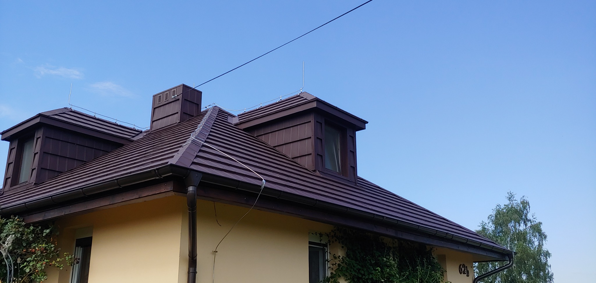 Dom z brązowym dachem z dachówki metalowej, widoczne okna dachowe, rynny i instalacja odgromowa, elewacja w kolorze kremowym, fragment zieleni pnącej się po ścianie.