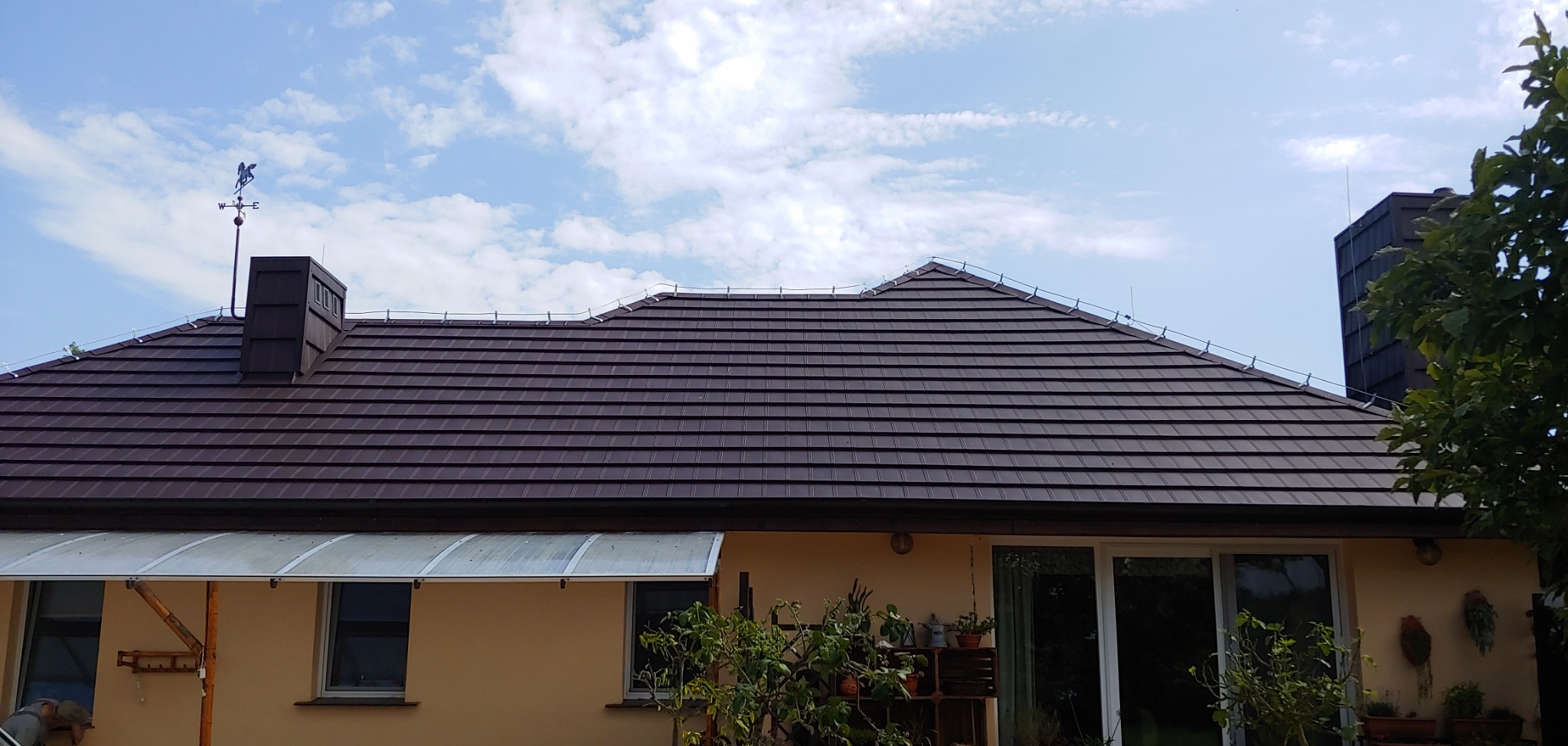 Brązowy dach z metalowej dachówki z kominem i wiatrowskazem w kształcie konia na tle błękitnego nieba z chmurami.